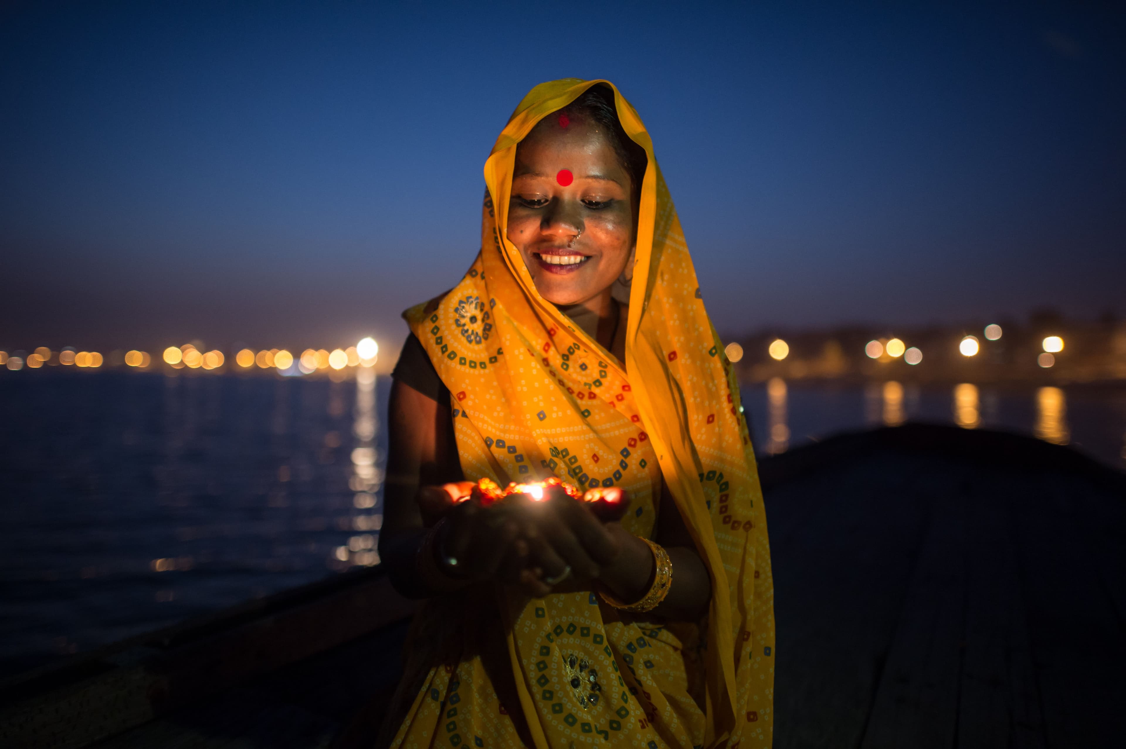 Voyage bien-être – Femme indienne en sari jaune tenant une lampe allumée au bord du Gange au crépuscule, lors d’un rituel lumineux empreint de spiritualité.